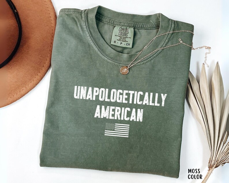 Może przedstawiać: Oliwkowo-zielona koszulka z białym napisem "UNAPOLOGETICALLY AMERICAN" i małą amerykańską flagą. Złoty naszyjnik z wisiorkiem spoczywa na koszulce. Koszulka jest złożona i wyeksponowana na białej powierzchni.