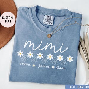 May include: A blue jean-colored t-shirt with a white daisy design and the text "mimi" above the daisies. Below the daisies, the text "emma james liam" is printed.