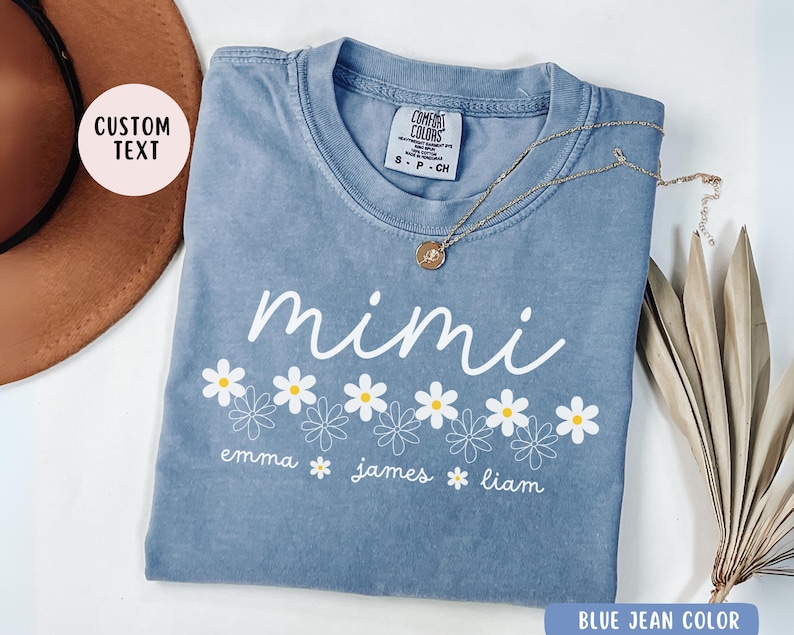 May include: A blue jean-colored t-shirt with a white daisy design and the text "mimi" above the daisies. Below the daisies, the text "emma james liam" is printed.