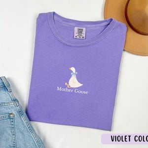 May include: A purple t-shirt featuring a white goose illustration and the text "Mother Goose" printed on the front. The t-shirt is made from a soft, comfortable cotton blend.