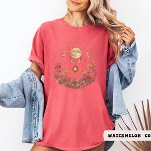 Może przedstawiać: Czerwony T-shirt z grafiką przedstawiającą księżyc, gwiazdy i kwiaty. Tekst "WATERMELON COLOR" jest widoczny w prawym dolnym rogu obrazu.