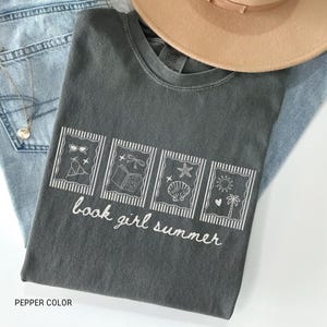 May include: A dark gray t-shirt with the text "book girl summer" and four illustrated rectangles. The illustrations include sunglasses, a tent, a book, a seashell, a sun, and a palm tree. The shirt is paired with blue jeans and a tan hat.