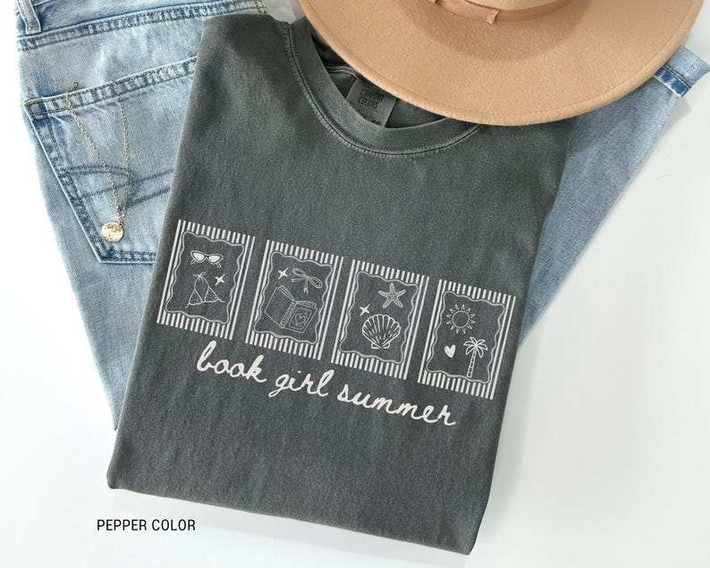 May include: A dark gray t-shirt with the text "book girl summer" and four illustrated rectangles. The illustrations include sunglasses, a tent, a book, a seashell, a sun, and a palm tree. The shirt is paired with blue jeans and a tan hat.