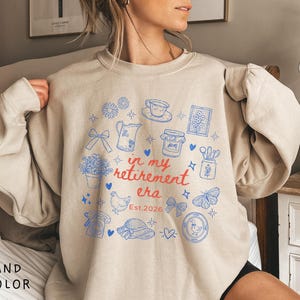 K&ouml;nnte beinhalten: Sandfarbenes Sweatshirt mit blau-roter Grafik. Die Grafik zeigt Illustrationen wie Blumen, eine Teekanne und einen Schmetterling sowie den Text "in my retirement era Est.2026".