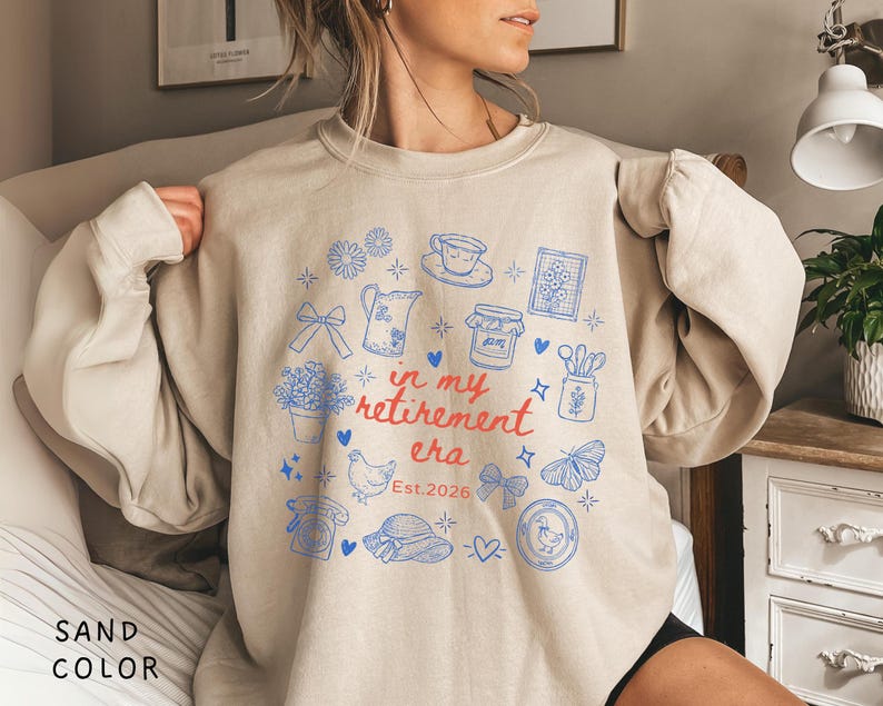 K&ouml;nnte beinhalten: Sandfarbenes Sweatshirt mit blau-roter Grafik. Die Grafik zeigt Illustrationen wie Blumen, eine Teekanne und einen Schmetterling sowie den Text "in my retirement era Est.2026".