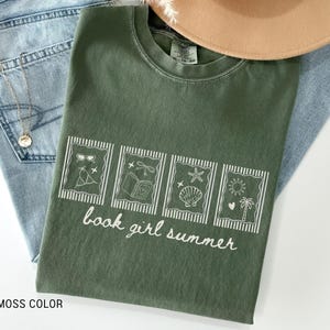 May include: A moss-colored t-shirt with a white graphic that reads "book girl summer." The graphic features four framed images of summer-themed items. The shirt is laid on top of blue jeans and a tan hat.