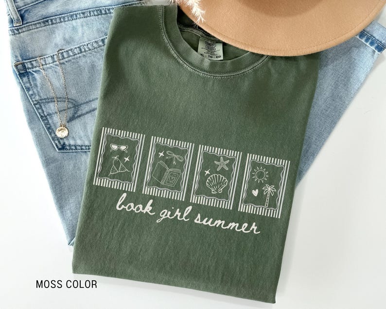 May include: A moss-colored t-shirt with a white graphic that reads "book girl summer." The graphic features four framed images of summer-themed items. The shirt is laid on top of blue jeans and a tan hat.