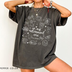K&ouml;nnte beinhalten: Oversized T-Shirt in Pfefferfarbe mit wei&szlig;em Grafikdesign. Das Design enth&auml;lt den Text "in my retirement era Est. 2026" sowie verschiedene Illustrationen wie Blumen, eine Teekanne, eine Tasse und einen Schmetterling.