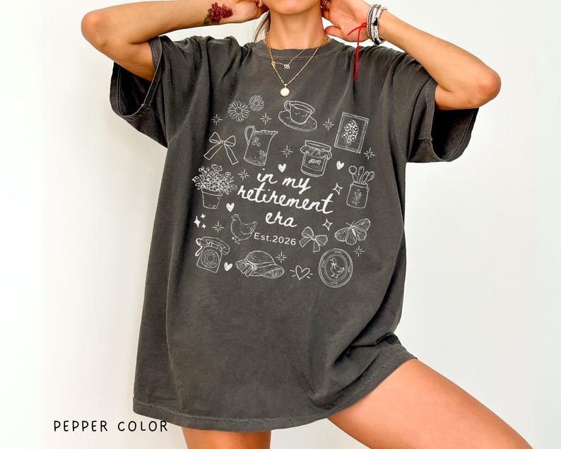K&ouml;nnte beinhalten: Oversized T-Shirt in Pfefferfarbe mit wei&szlig;em Grafikdesign. Das Design enth&auml;lt den Text "in my retirement era Est. 2026" sowie verschiedene Illustrationen wie Blumen, eine Teekanne, eine Tasse und einen Schmetterling.