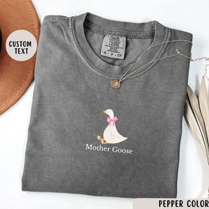 May include: A grey t-shirt featuring a white graphic of a goose with a pink bow. The text "Mother Goose" is printed below the goose. The shirt is made from a soft, comfortable material.