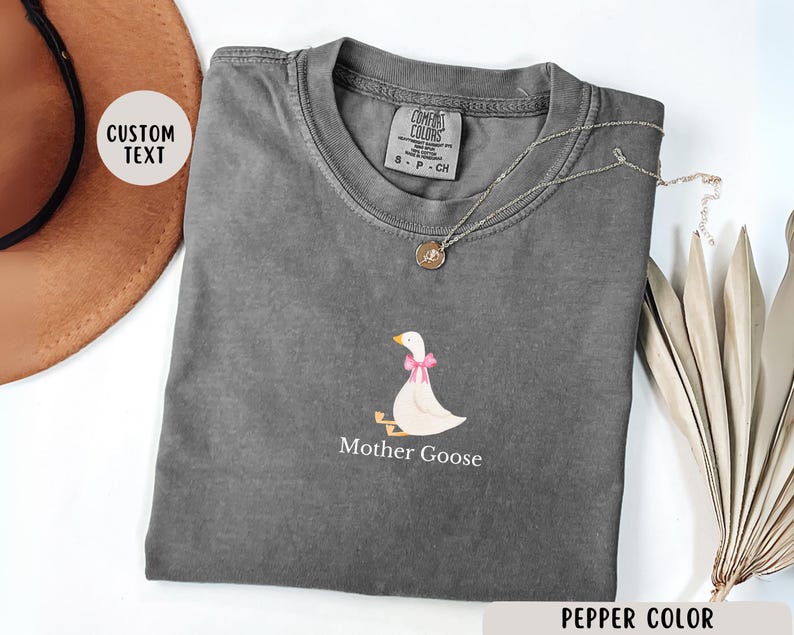 May include: A grey t-shirt featuring a white graphic of a goose with a pink bow. The text "Mother Goose" is printed below the goose. The shirt is made from a soft, comfortable material.