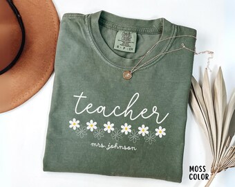 May include: A moss green t-shirt with the word "teacher" in white script, decorated with white and yellow daisy graphics. The name "mrs. johnson" is printed below the daisies. A gold necklace and a brown hat are visible.