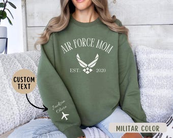 May include: Green sweatshirt with the text "Air Force Mom Est. 2020" and a white Air Force logo. The sweatshirt has a white airplane graphic on the sleeve and the text "Jackson Olivia" on the sleeve.