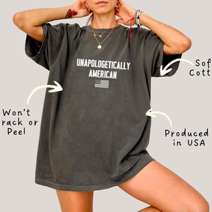 Może przedstawiać: Ciemnoszara koszulka z napisem "UNAPOLOGETICALLY AMERICAN" i małą amerykańską flagą. Koszulka wykonana jest z miękkiej bawełny i wyprodukowana w USA. Nadruk jest odporny na pękanie i łuszczenie się.