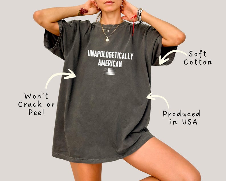 Może przedstawiać: Ciemnoszara koszulka z napisem "UNAPOLOGETICALLY AMERICAN" i małą amerykańską flagą. Koszulka wykonana jest z miękkiej bawełny i wyprodukowana w USA. Nadruk jest odporny na pękanie i łuszczenie się.