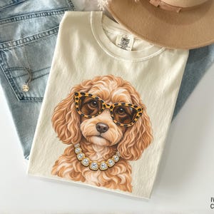 May include: An ivory t-shirt featuring a graphic illustration of a curly-haired dog wearing leopard print glasses and a square-cut crystal necklace. The shirt is layered with light blue denim jeans, a silver necklace, and a tan wide-brimmed hat.