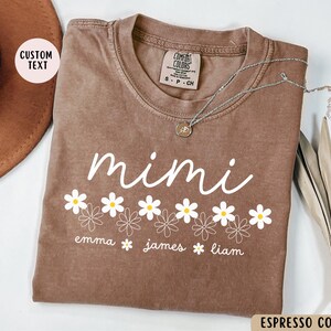 May include: Espresso brown t-shirt with the word "mimi" in white script and a row of white daisies with yellow centers. Below the daisies are the names "emma", "james", and "liam" in white text.