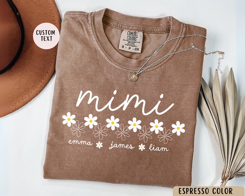 May include: Espresso brown t-shirt with the word "mimi" in white script and a row of white daisies with yellow centers. Below the daisies are the names "emma", "james", and "liam" in white text.