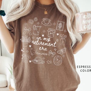 K&ouml;nnte beinhalten: Braunes T-Shirt mit wei&szlig;em Grafikdesign, das eine Tasse, Blumen und ein Huhn zeigt. Der Text "in my retirement era" und "Est. 2026" sind ebenfalls enthalten. Das Shirt hat eine lockere Passform. Die Farbe ist Espresso.