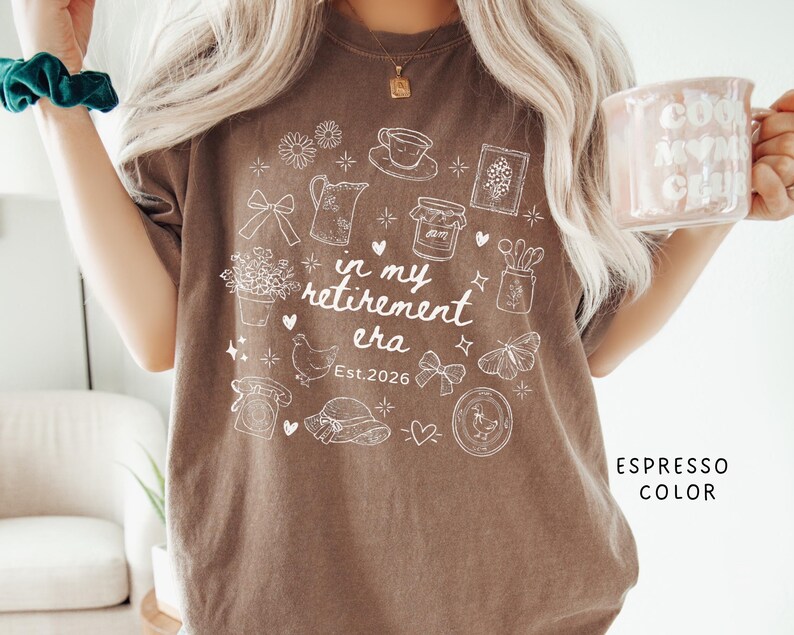 K&ouml;nnte beinhalten: Braunes T-Shirt mit wei&szlig;em Grafikdesign, das eine Tasse, Blumen und ein Huhn zeigt. Der Text "in my retirement era" und "Est. 2026" sind ebenfalls enthalten. Das Shirt hat eine lockere Passform. Die Farbe ist Espresso.