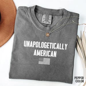 Może przedstawiać: Szara koszulka z białym napisem "UNAPOLOGETICALLY AMERICAN" i małą grafiką amerykańskiej flagi. Koszulka jest złożona i ma metkę Comfort Colors. Widoczny jest r&oacute;wnież złoty naszyjnik i brązowy kapelusz.