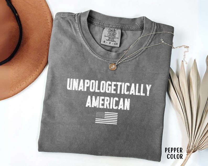 Może przedstawiać: Szara koszulka z białym napisem "UNAPOLOGETICALLY AMERICAN" i małą grafiką amerykańskiej flagi. Koszulka jest złożona i ma metkę Comfort Colors. Widoczny jest r&oacute;wnież złoty naszyjnik i brązowy kapelusz.