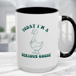 May include: White ceramic coffee mug with a black rim and handle. The mug has a green design of a goose wearing a suit and the text "Today I'm a Serious Goose".