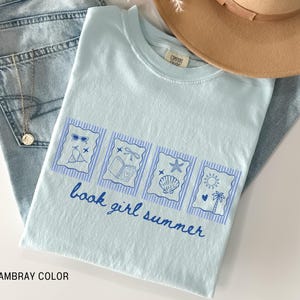 May include: Light blue t-shirt with a graphic design featuring sunglasses, a bikini, a book, a dragonfly, a starfish, a seashell, a sun, and a palm tree. The text "book girl summer" is written in blue script. The shirt is folded on top of blue jeans.
