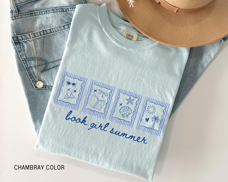 May include: Light blue t-shirt with a graphic design featuring sunglasses, a bikini, a book, a dragonfly, a starfish, a seashell, a sun, and a palm tree. The text "book girl summer" is written in blue script. The shirt is folded on top of blue jeans.