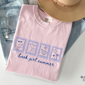 May include: A light pink t-shirt with a blue graphic that says "book girl summer." The graphic includes images of sunglasses, a book, a seashell, and a sun. The shirt is folded on top of blue jeans and a tan hat.