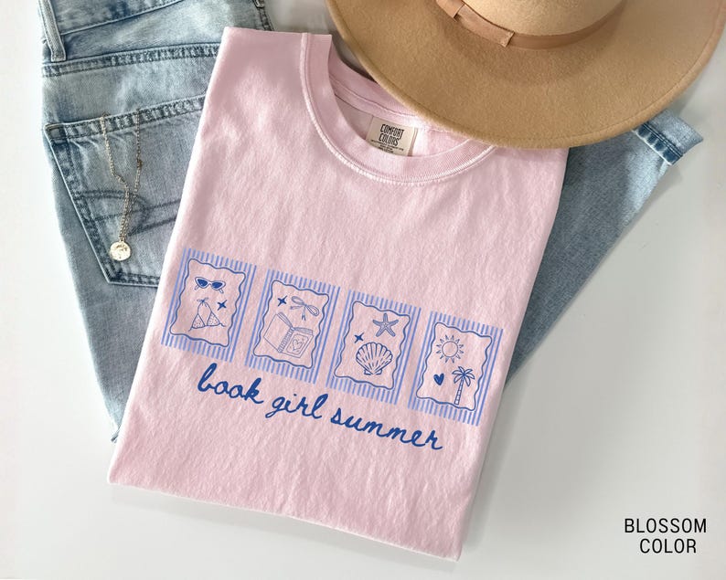 May include: A light pink t-shirt with a blue graphic that says "book girl summer." The graphic includes images of sunglasses, a book, a seashell, and a sun. The shirt is folded on top of blue jeans and a tan hat.