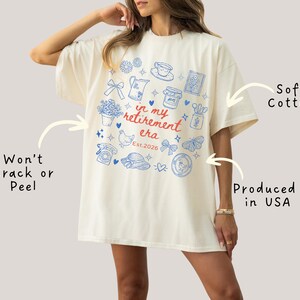 K&ouml;nnte beinhalten: Cremefarbenes &uuml;bergro&szlig;es T-Shirt mit blauem und rotem Grafikdesign. Das Design enth&auml;lt verschiedene Illustrationen und den Text "in my retirement era Est.2026". Das Shirt besteht aus weicher Baumwolle und wird in den USA hergestellt.