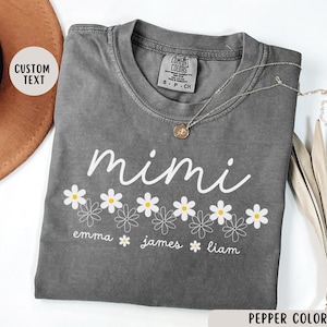 May include: Gray t-shirt with the word "mimi" in white script above a row of white daisies with yellow centers. Below the daisies are the names "emma", "james", and "liam" in white text.