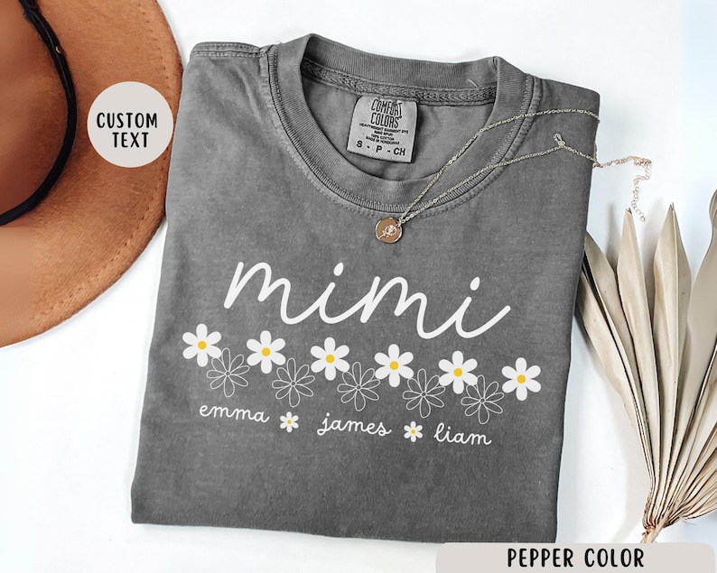 May include: Gray t-shirt with the word "mimi" in white script above a row of white daisies with yellow centers. Below the daisies are the names "emma", "james", and "liam" in white text.