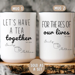 May include: Two white ceramic mugs with black handles and black text. The first mug says "Let's have a tea together" with the names "Emily" and "Mathew" and the date "EST. 2025". The second mug says "For the res of our lives" with the names "Emily" and "Mathew" and the date "EST. 2025".