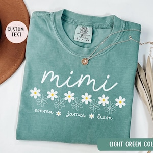 May include: Light green t-shirt with the word "mimi" in white lettering and a row of white daisies with yellow centers. The text "emma james liam" is written below the daisies.