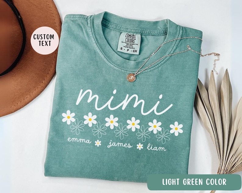 May include: Light green t-shirt with the word "mimi" in white lettering and a row of white daisies with yellow centers. The text "emma james liam" is written below the daisies.