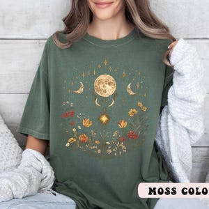 Może przedstawiać: Zielony t-shirt w kolorze mchu z wzorem księżyca i kwiat&oacute;w. Księżyc jest otoczony gwiazdami i p&oacute;łksiężycami. Wz&oacute;r kwiatowy zawiera kwiaty, liście i winorośl.