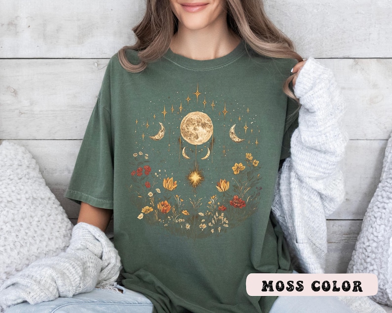 Może przedstawiać: Zielony t-shirt w kolorze mchu z wzorem księżyca i kwiat&oacute;w. Księżyc jest otoczony gwiazdami i p&oacute;łksiężycami. Wz&oacute;r kwiatowy zawiera kwiaty, liście i winorośl.