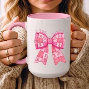 May include: White ceramic mug with a pink interior and handle. The mug features a large pink ribbon design with the text "In October We Wear Pink". The design is a patchwork of pink ribbons and squares.