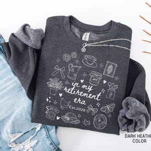 K&ouml;nnte beinhalten: Dunkelgraues Sweatshirt mit wei&szlig;en Illustrationen und dem Text "in my retirement era Est. 2026". Eine silberne Halskette liegt &uuml;ber dem Sweatshirt. Das Sweatshirt liegt gefaltet neben einer blauen Jeans.