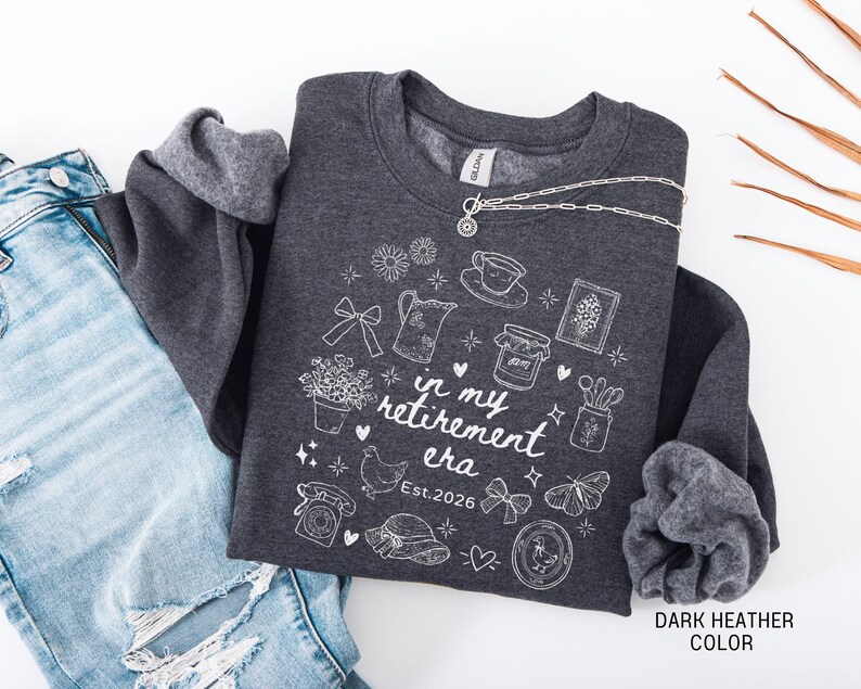 K&ouml;nnte beinhalten: Dunkelgraues Sweatshirt mit wei&szlig;en Illustrationen und dem Text "in my retirement era Est. 2026". Eine silberne Halskette liegt &uuml;ber dem Sweatshirt. Das Sweatshirt liegt gefaltet neben einer blauen Jeans.