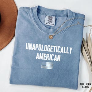 Może przedstawiać: Niebieska koszulka w kolorze dżinsu z białym napisem "UNAPOLOGETICALLY AMERICAN" i amerykańską flagą. Widoczny jest r&oacute;wnież złoty naszyjnik i brązowy kapelusz.
