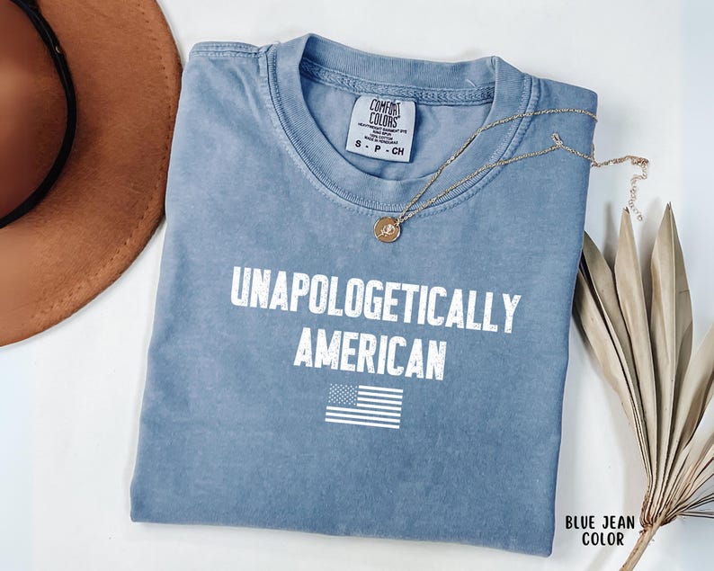 Może przedstawiać: Niebieska koszulka w kolorze dżinsu z białym napisem "UNAPOLOGETICALLY AMERICAN" i amerykańską flagą. Widoczny jest r&oacute;wnież złoty naszyjnik i brązowy kapelusz.