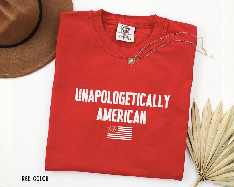 Może przedstawiać: Czerwony t-shirt z okrągłym dekoltem z białym napisem "UNAPOLOGETICALLY AMERICAN" i małą grafiką amerykańskiej flagi. Widoczny jest r&oacute;wnież złoty naszyjnik z wisiorkiem i brązowy kapelusz.