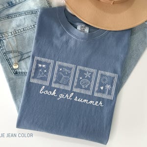 May include: A blue jean-colored t-shirt with the text "book girl summer" and four white framed illustrations. The illustrations depict sunglasses, a bow, a seashell, and a sun with a palm tree. A light brown hat and blue jeans are also in the image.