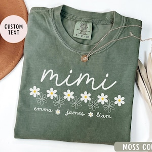 May include: A green t-shirt with the word "mimi" in white script and a row of white daisies with yellow centers. Below the daisies are the names "emma", "james", and "liam" in white text. The shirt is a comfortable color and has a relaxed fit.