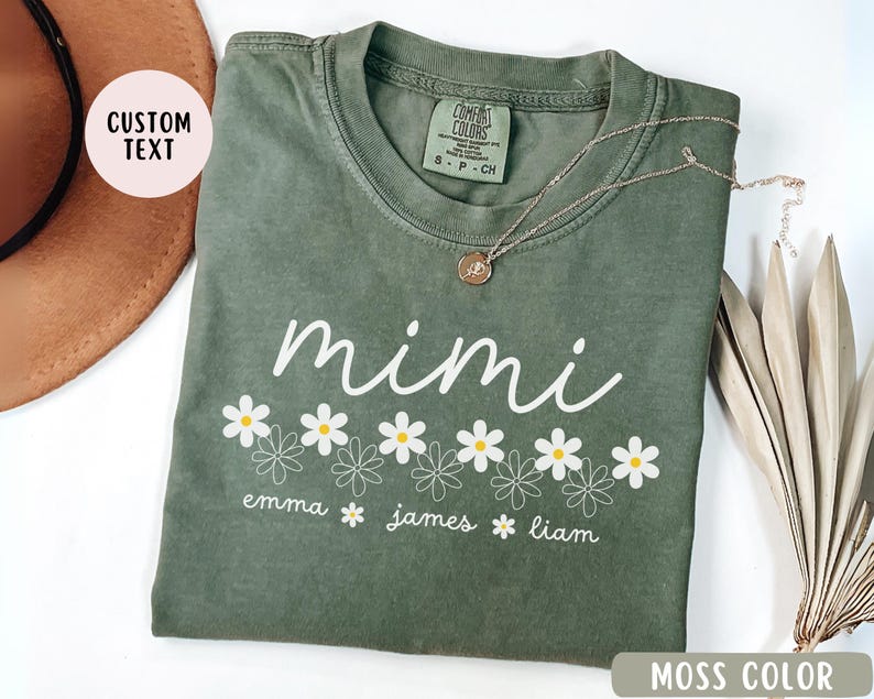 May include: A green t-shirt with the word "mimi" in white script and a row of white daisies with yellow centers. Below the daisies are the names "emma", "james", and "liam" in white text. The shirt is a comfortable color and has a relaxed fit.