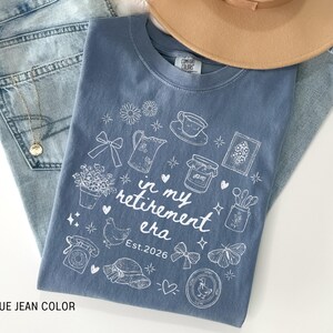 K&ouml;nnte beinhalten: Ein blaues Jeans-T-Shirt mit einem wei&szlig;en Aufdruck, der "in my retirement era Est. 2026" lautet. Das Shirt ist mit Illustrationen von Blumen, einer Teekanne und einem Schmetterling verziert. Ein beiger Hut und Jeans sind ebenfalls im Bild.