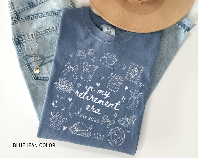 K&ouml;nnte beinhalten: Ein blaues Jeans-T-Shirt mit einem wei&szlig;en Aufdruck, der "in my retirement era Est. 2026" lautet. Das Shirt ist mit Illustrationen von Blumen, einer Teekanne und einem Schmetterling verziert. Ein beiger Hut und Jeans sind ebenfalls im Bild.